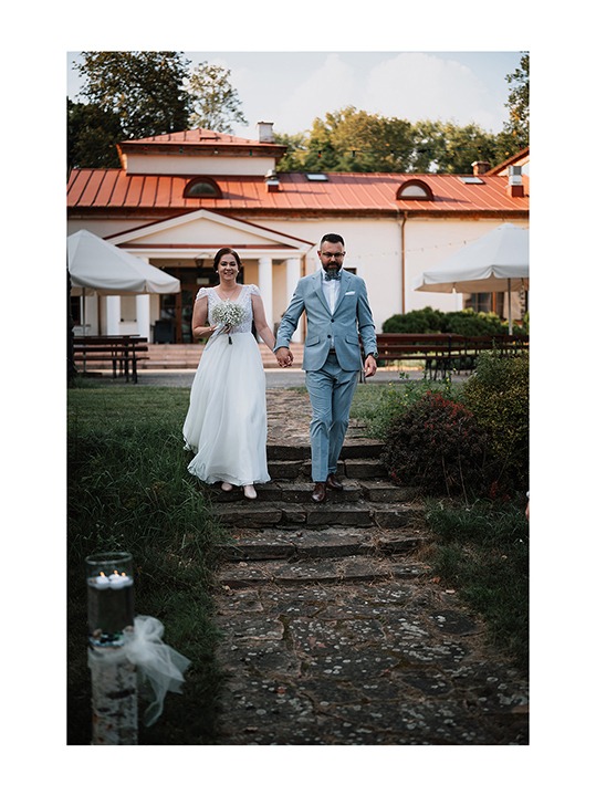 Para młoda w jasnych strojach spaceruje po kamiennych schodach na tle budynku z czerwoną dachówką. Panna młoda trzyma bukiet.