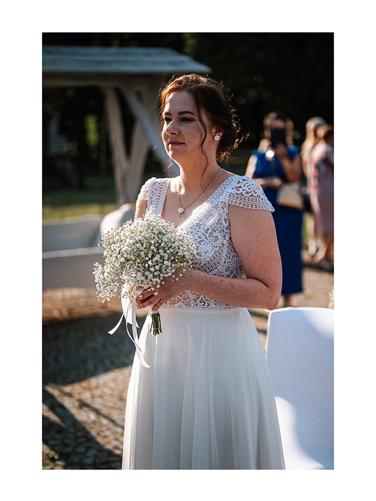 Panna młoda w koronkowej sukni trzyma bukiet gipsówki. W tle goście weselni i biała altana. Delikatne światło podkreśla uroczysty charakter wydarzenia.