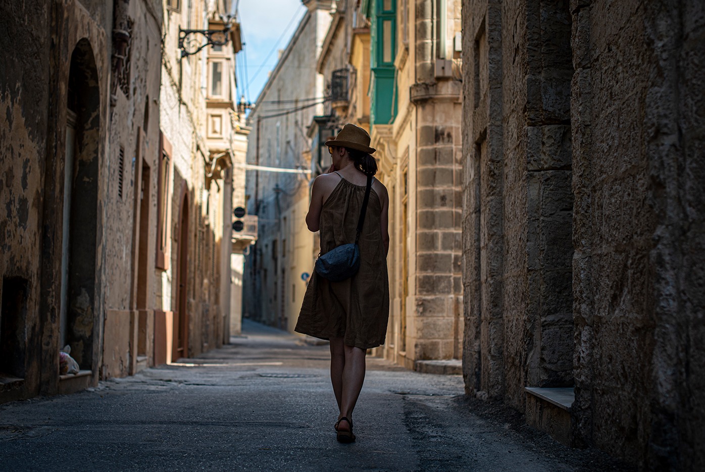 Kobieta w brązowej sukience i kapeluszu idzie wąską, zacienioną uliczką w starym mieście, z kamiennymi budynkami po obu stronach i lekkim światłem na końcu ulicy.