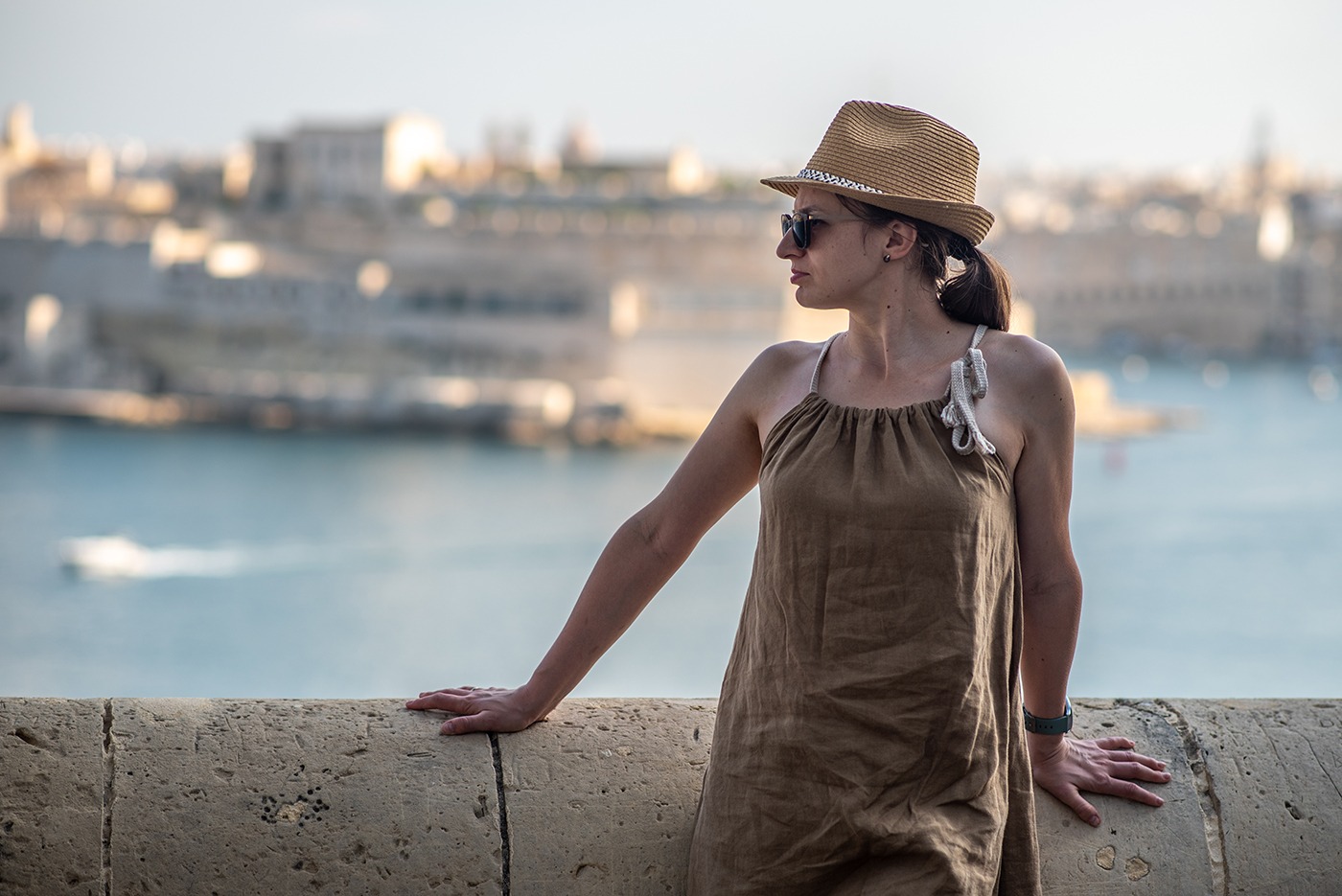 Kobieta w brązowej sukience i słomkowym kapeluszu, oparta o kamienny mur z widokiem na miasto i morze w tle, w okularach przeciwsłonecznych.