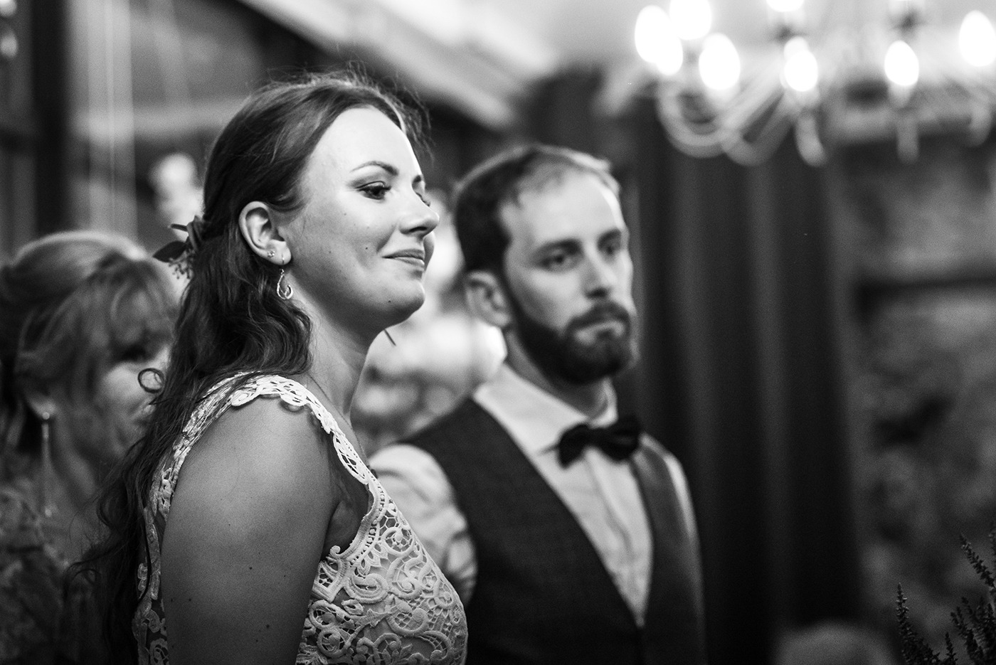 Czarno-białe zdjęcie z wesela: uśmiechnięta panna młoda w koronkowej sukni, obok pan młody w muszce, w tle rozmyte światła żyrandola.