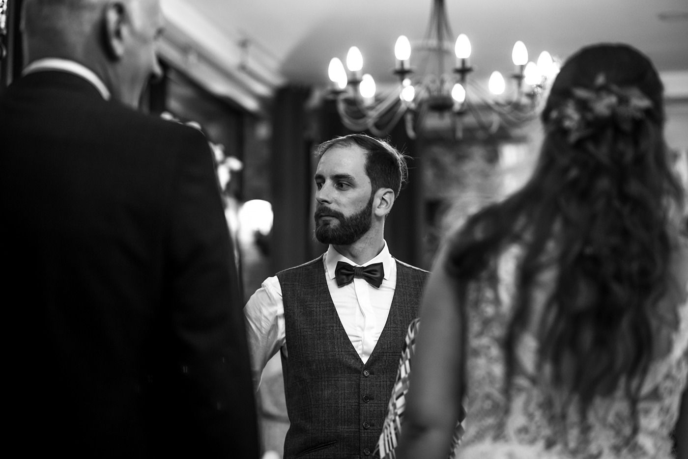 Czarno-białe zdjęcie mężczyzny z brodą w muszce i kraciastej kamizelce, stojącego w pomieszczeniu z żyrandolem, widzianego z perspektywy osoby stojącej naprzeciwko.