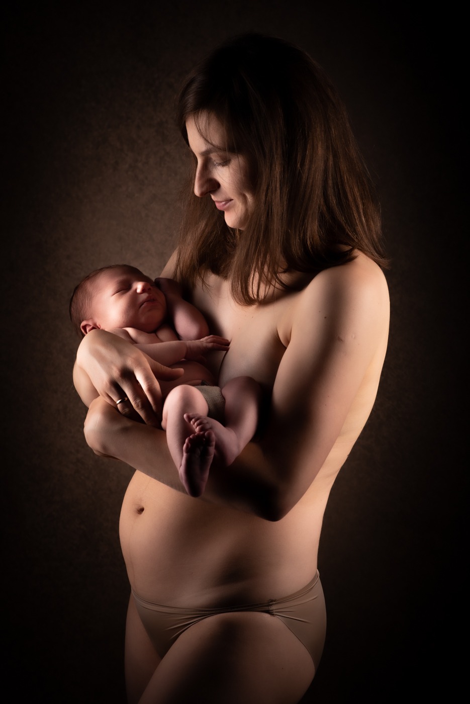 Portret matki trzymającej śpiącego noworodka na tle ciemnego, teksturowanego tła, z bocznym oświetleniem podkreślającym kontury ciała.