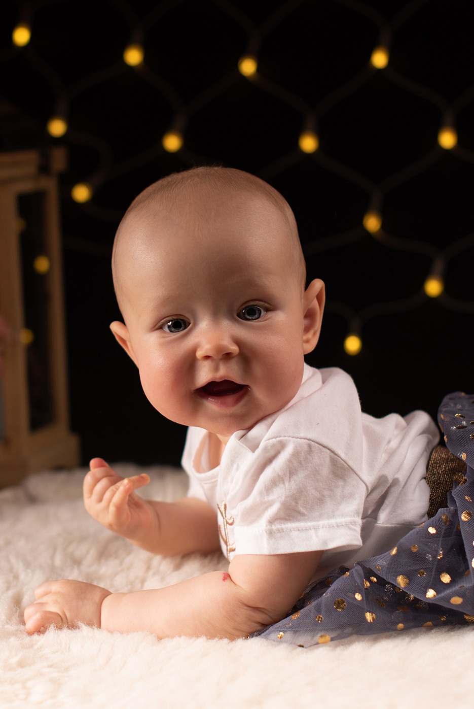Urocze niemowlę leży na brzuszku na puszystym dywanie, ubrane w białą koszulkę i tiulową spódniczkę w złote kropki, w tle ciemne tło z rozproszonymi światełkami.
