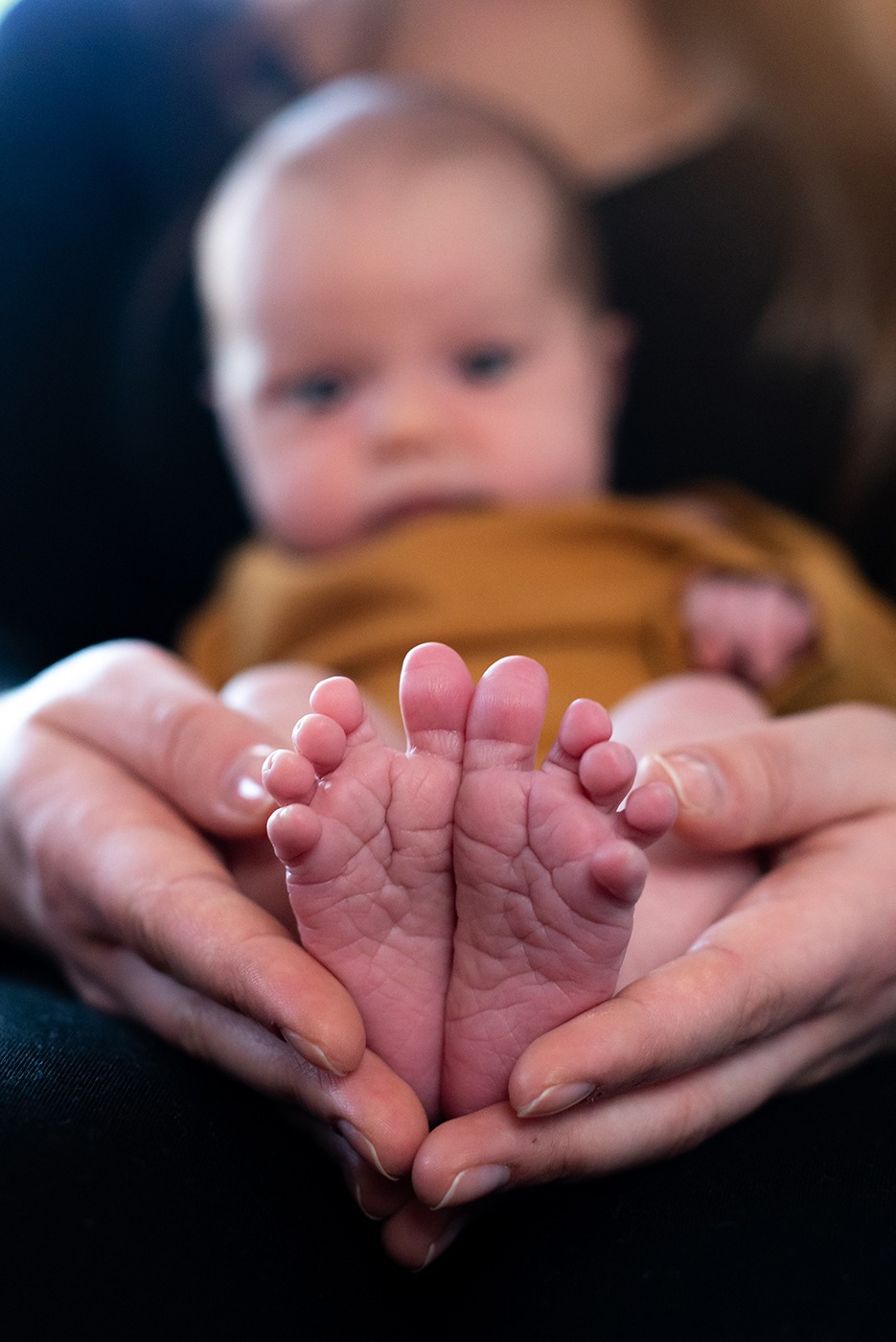 Drobne stópki noworodka trzymane w dłoniach rodzica, z delikatnymi fałdkami skóry i widocznymi paznokciami, na rozmytym tle twarzy dziecka.