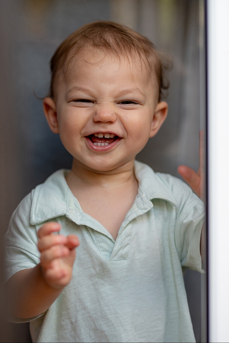 Rozesmiany roczny chłopiec w jasnej koszulce polo, z widocznymi pierwszymi ząbkami, pozuje w kadrze bliskim, z lekko rozmazanym tłem.