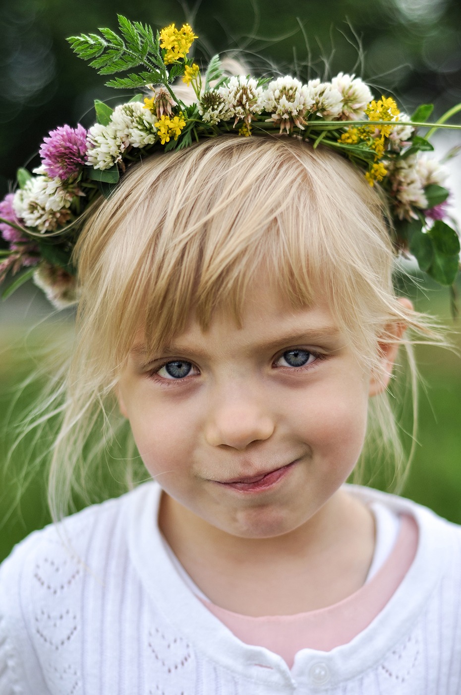Portret uśmiechniętej dziewczynki z blond włosami i niebieskimi oczami, w wianku z polnych kwiatów na głowie, ubrana w biały sweter