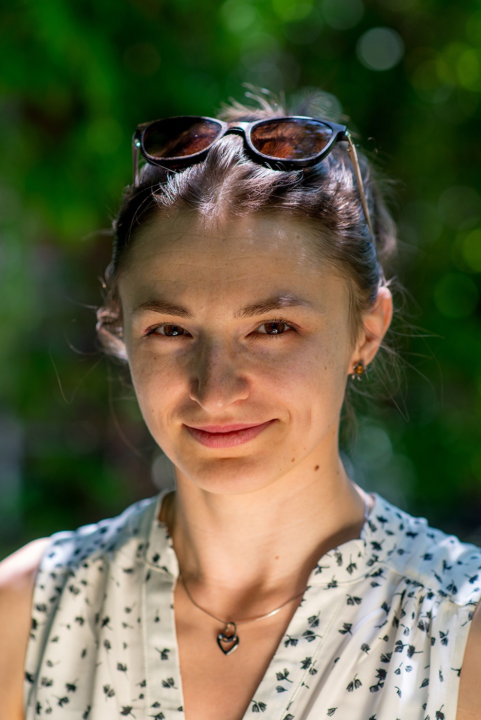 Portret uśmiechniętej kobiety z okularami na głowie, na tle rozmytej zieleni, w bluzce w drobne kwiatki i z naszyjnikiem w kształcie serca.