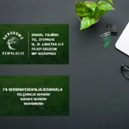 Dwie wizytówki firmy ogrodniczej 'Ogrodowe Rewolucje' na ciemnym biurku z laptopem, spinaczami, okularami i małą rośliną doniczkową.