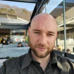 Portret mężczyzny z brodą na tle przeszklonej konstrukcji z widokiem na drzewa i osoby w oddali. Ubrany w ciemną koszulę w drobny wzór.