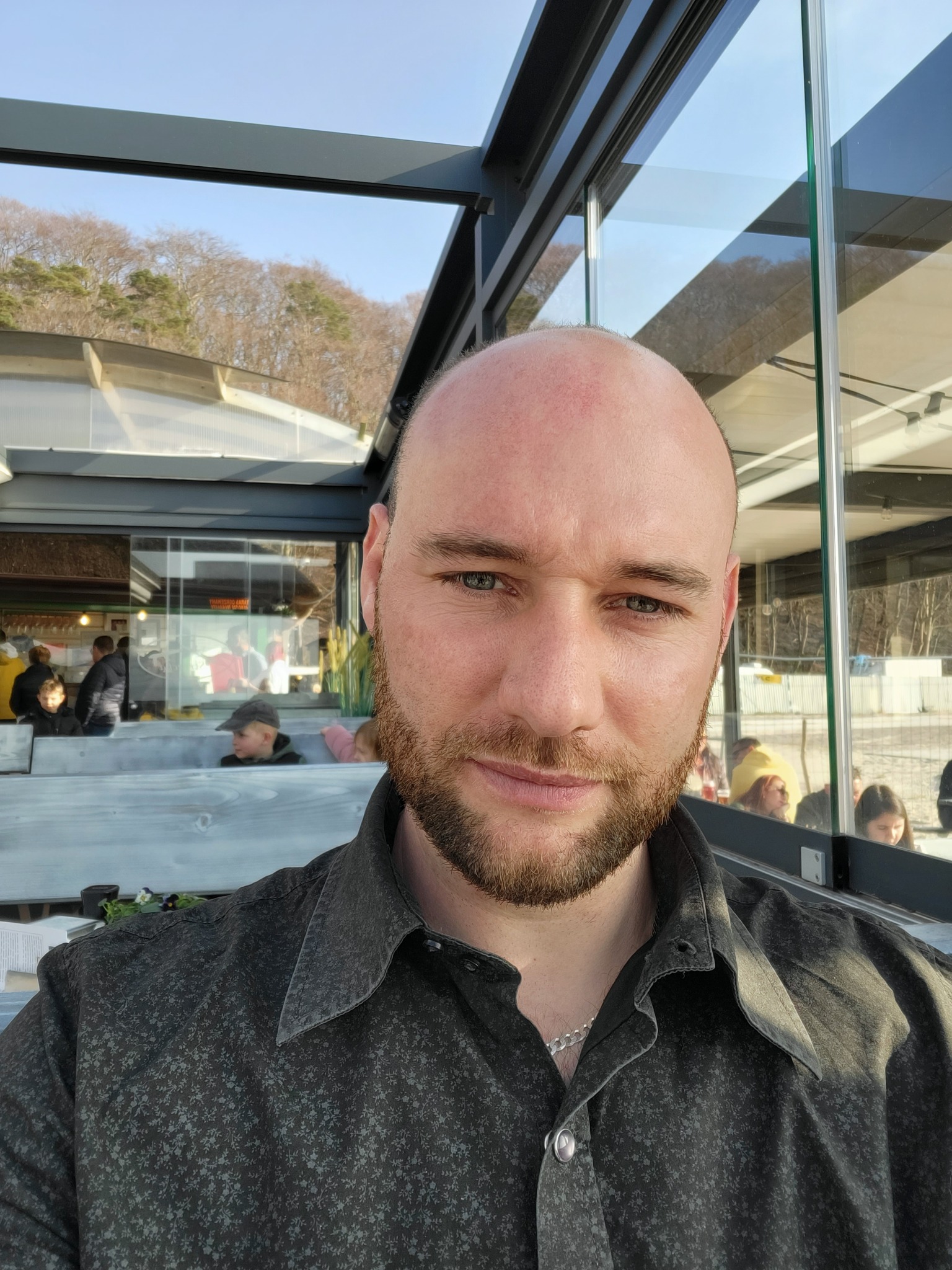 Portret mężczyzny z brodą na tle przeszklonej konstrukcji z widokiem na drzewa i osoby w oddali. Ubrany w ciemną koszulę w drobny wzór.