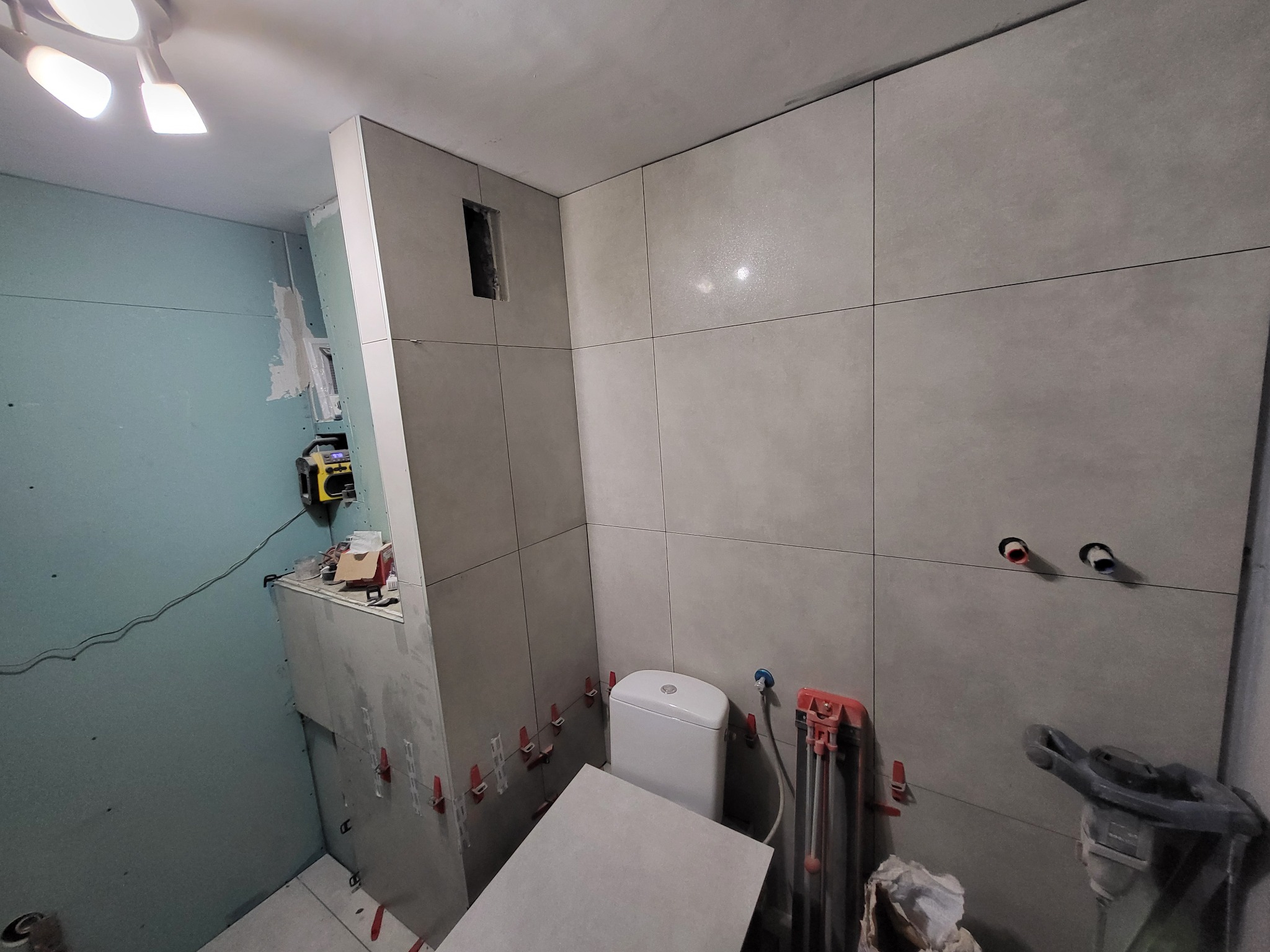 Wnętrze łazienki w trakcie wykańczania: szare płytki na ścianach, częściowo obudowana toaleta, narzędzia glazurnicze i płyta na podłodze. Widoczne elementy instalacji.