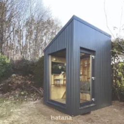 Nowoczesna, mała, szara konstrukcja z dużymi oknami, ustawiona w ogrodzie, z widocznym drewnianym wnętrzem i logo firmy na dole.