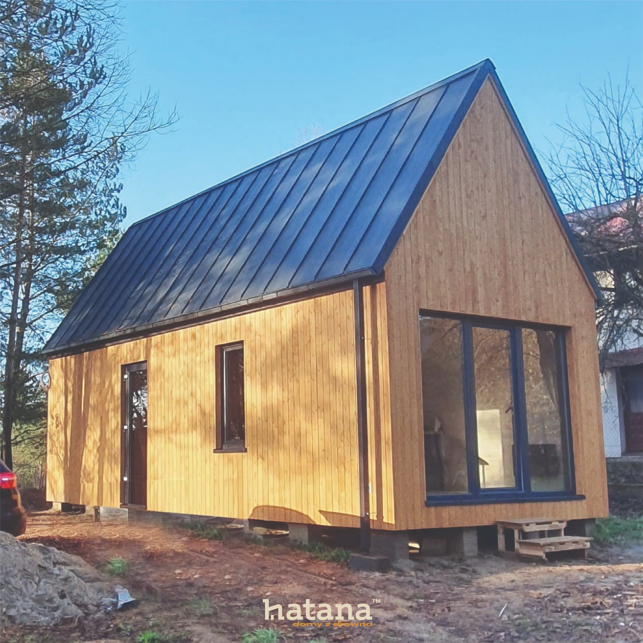 Nowoczesny, parterowy budynek z jasnego drewna z ciemnym, metalowym dachem, widoczne okna z ciemnymi ramami i drewniane schodki wejściowe, otoczony drzewami.