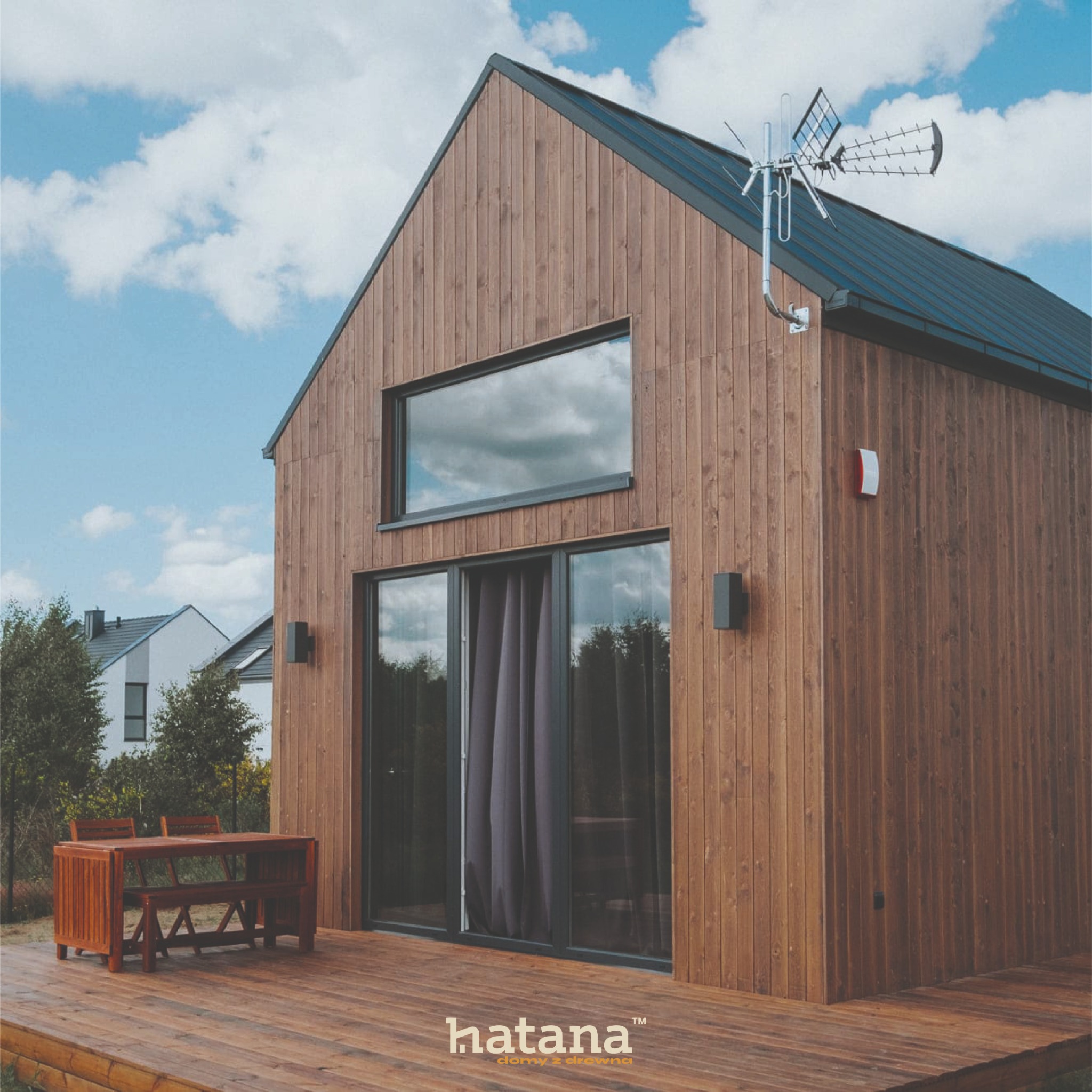 Nowoczesny dom szkieletowy z drewnianą elewacją, dużym oknem i tarasem z kompletem mebli ogrodowych, antena telewizyjna na dachu.