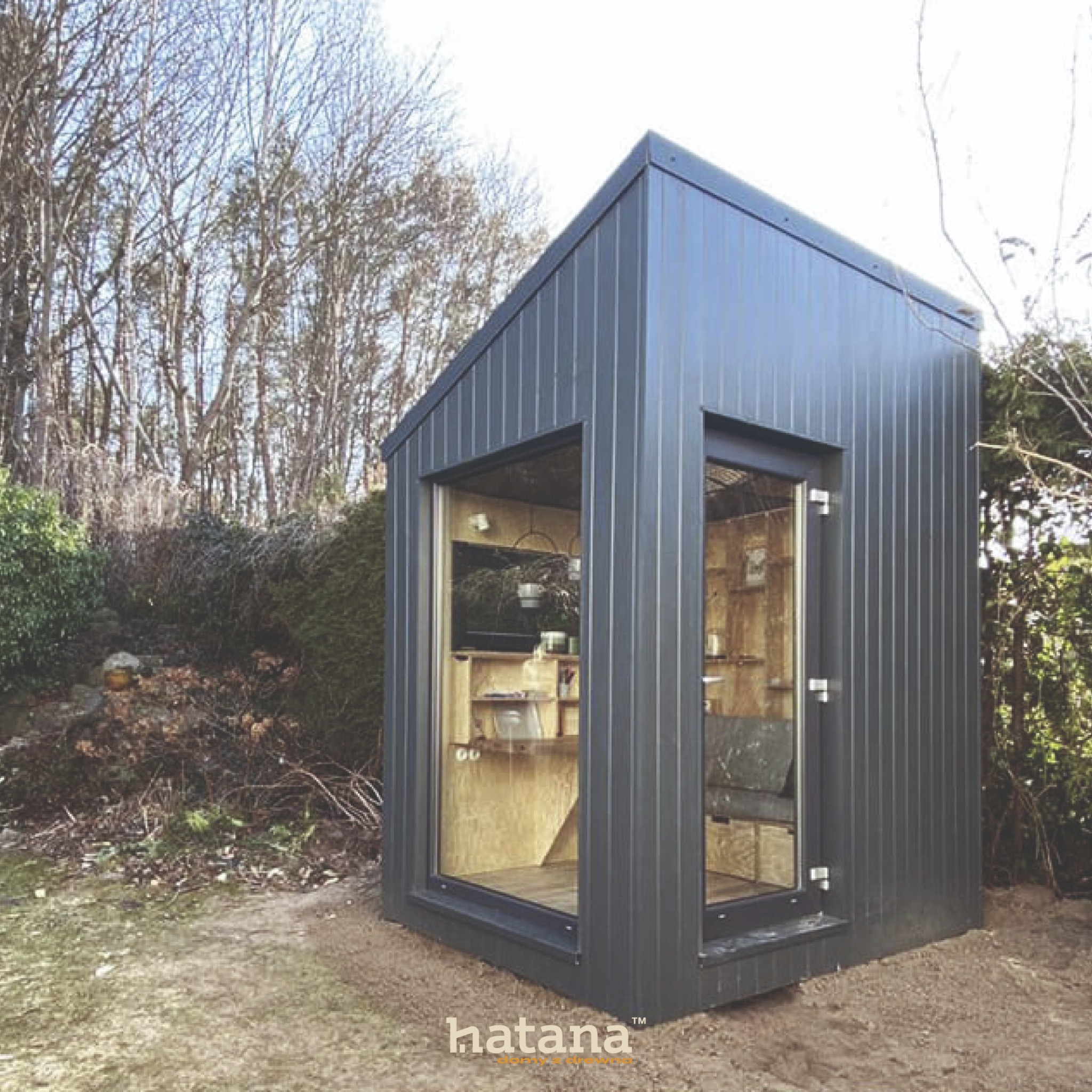 Nowoczesna, mała, szara konstrukcja z dużymi oknami, ustawiona w ogrodzie, z widocznym drewnianym wnętrzem i logo firmy na dole.