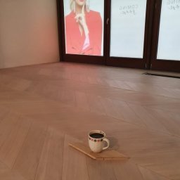 Kamil Maliszewski - Jasny parkiet ułożony we wzór jodełki, na którym stoi kubek z kawą na małej drewnianej deseczce. W tle duże okna z reklamą.