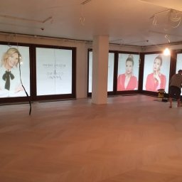 Kamil Maliszewski - Układanie jasnego parkietu w jodełkę w przestronnym wnętrzu z dużymi oknami z reklamami, widoczny pracownik przy pracy i wiszące kable pod sufitem.