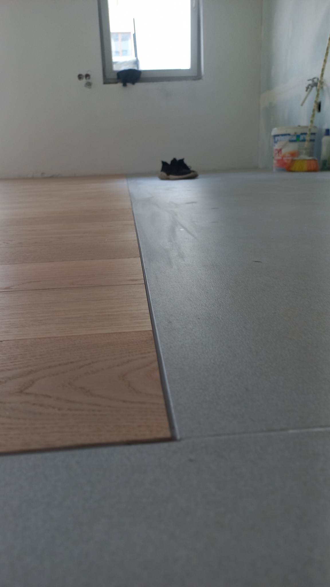 Fragment podłogi z widocznym połączeniem ułożonego parkietu z szarą wylewką samopoziomującą, w tle okno i pozostawione obuwie robocze.