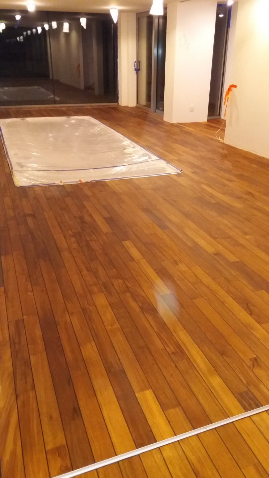 Świeżo polakierowany, lśniący parkiet z egzotycznego drewna, zabezpieczony folią ochronną na części powierzchni, w jasnym, nowoczesnym wnętrzu z dużymi oknami.