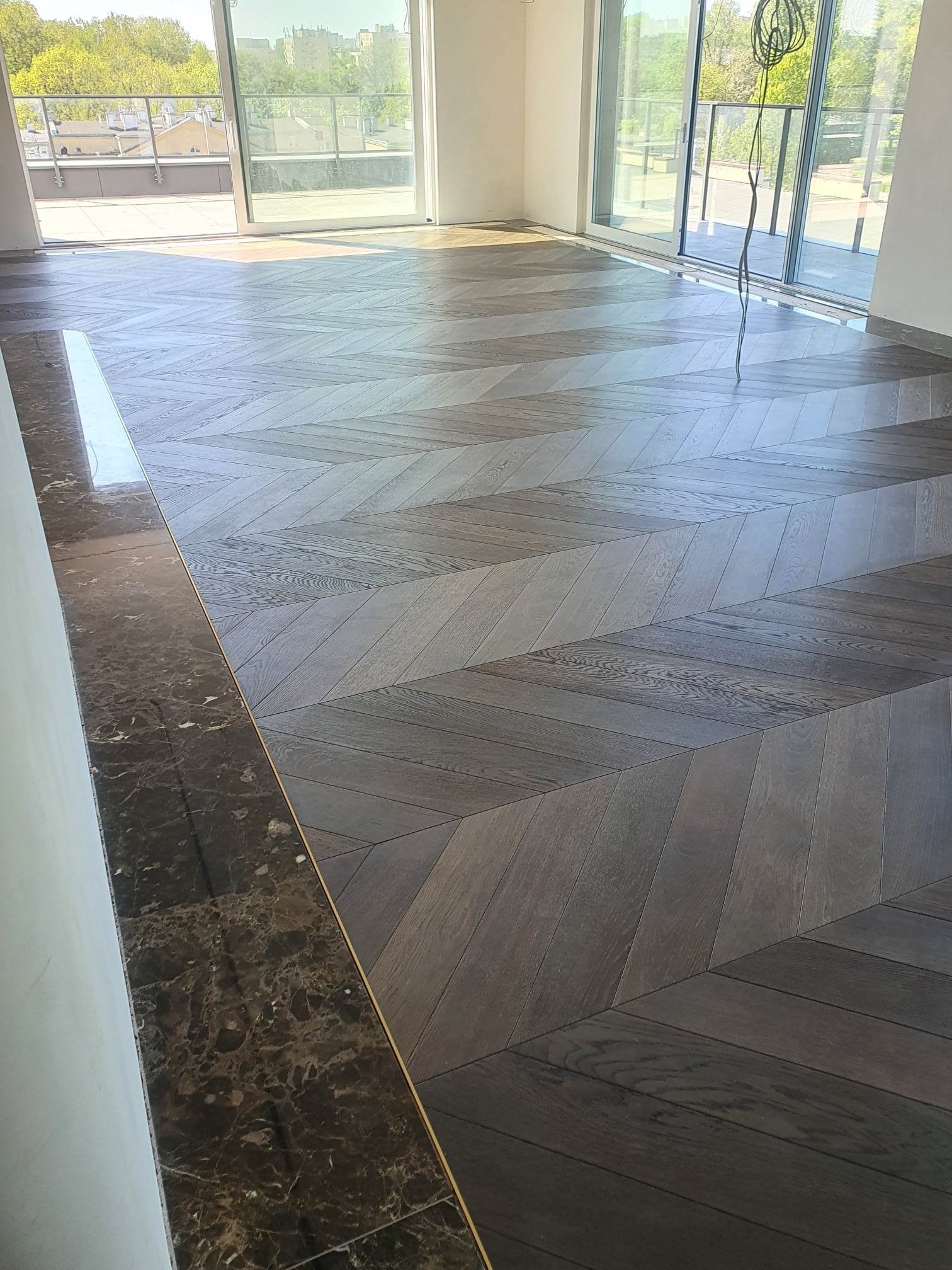 Nowo ułożony parkiet w jodełkę francuską z ciemnego drewna, wykończony marmurową listwą przypodłogową w nowoczesnym, jasnym wnętrzu z dużymi oknami balkonowymi.