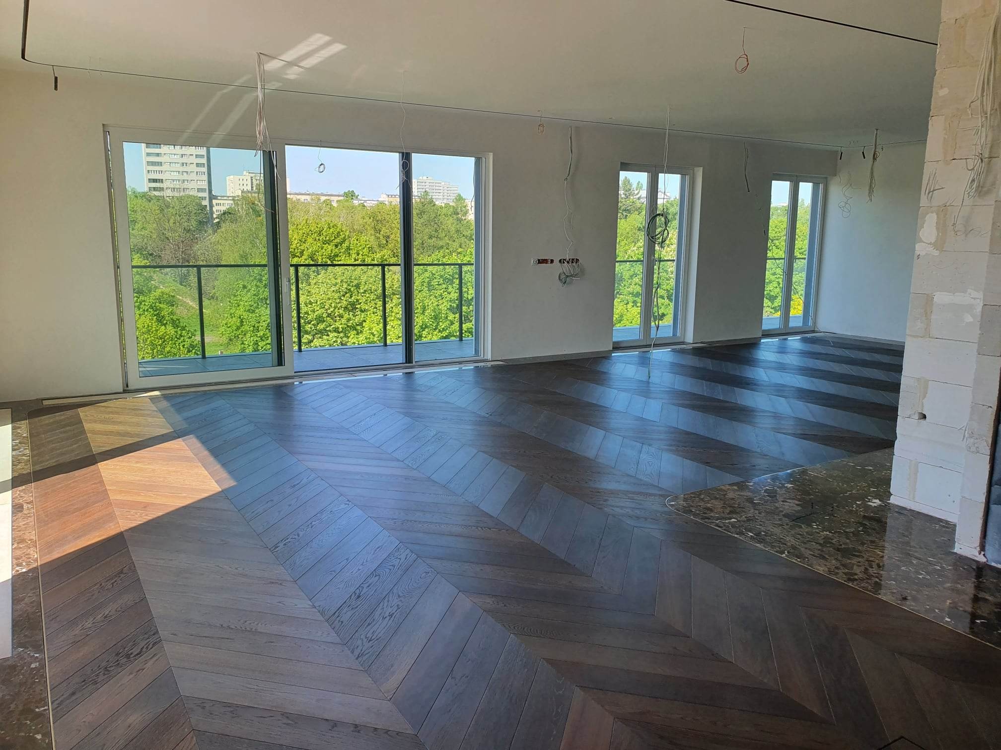 Nowoczesny salon z ułożonym parkietem w jodełkę, widok na zieleń za oknami, surowy stan wykończenia ścian i sufitu.