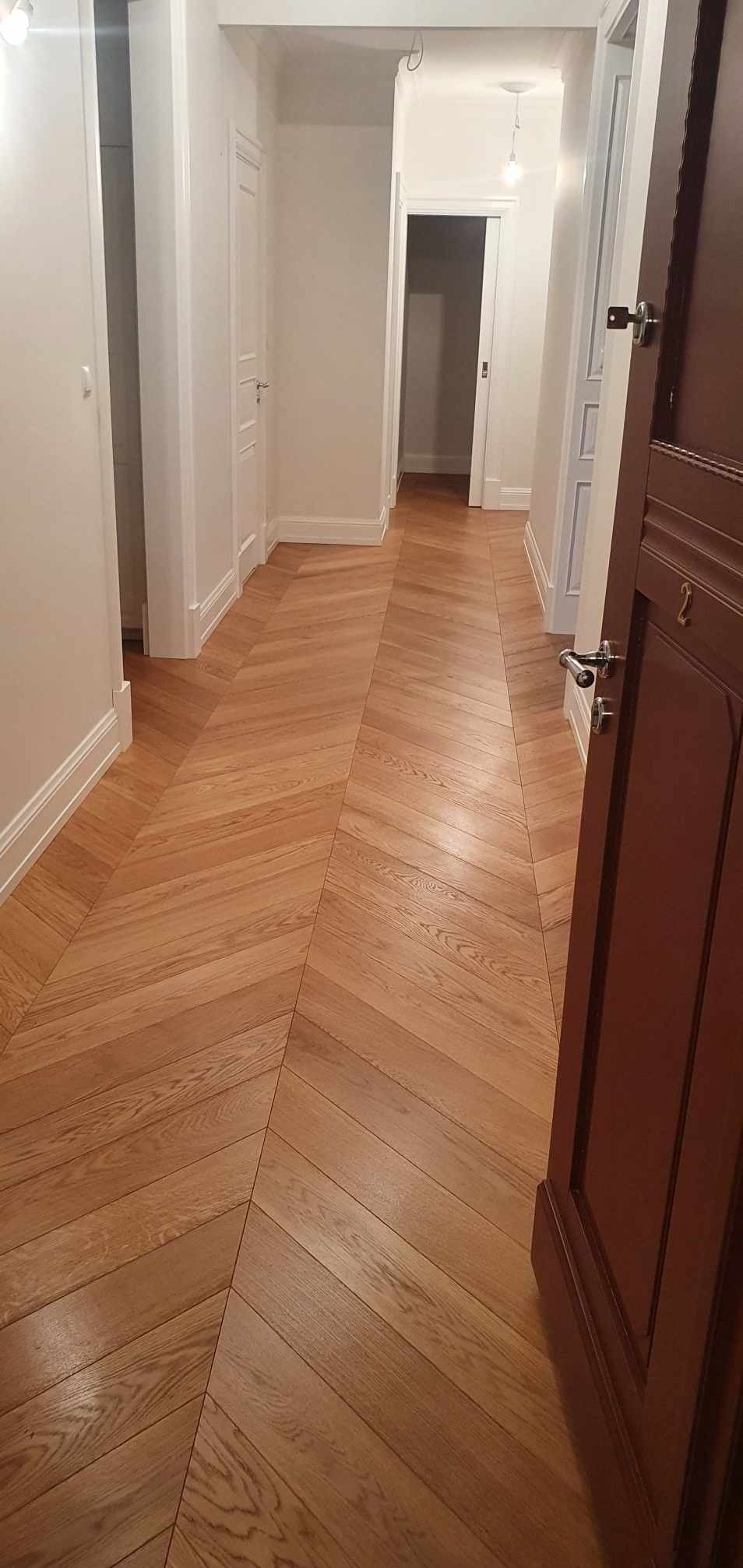 Korytarz z parkietem ułożonym we wzór jodełki francuskiej; widoczny fragment brązowych drzwi i białe ściany z listwami przypodłogowymi.