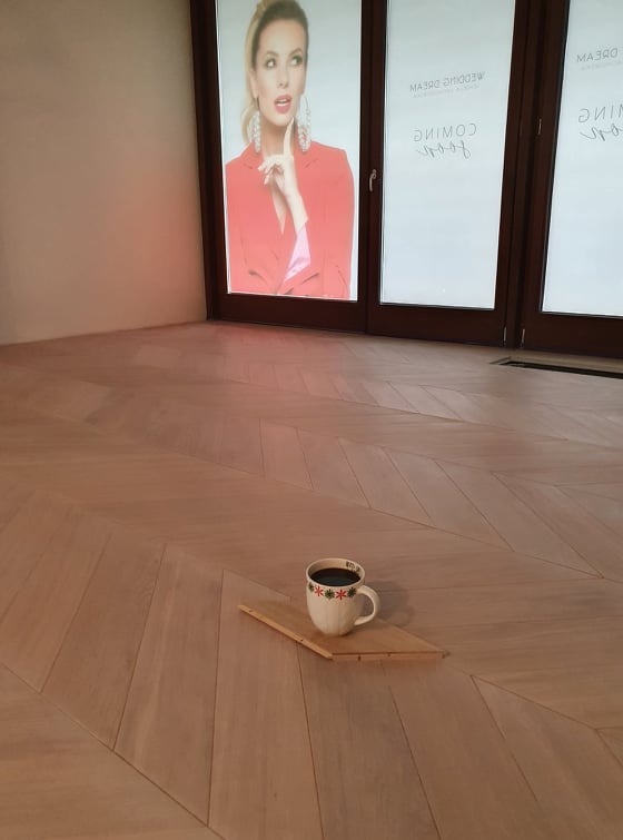 Jasny parkiet ułożony we wzór jodełki, na którym stoi kubek z kawą na małej drewnianej deseczce. W tle duże okna z reklamą.