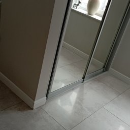 Magda Muszyńska - Fragment szafy przesuwnej z lustrami, odbijającej okno z kwiatami w wazonie i figurką psa, na tle jasnej podłogi z płytek.