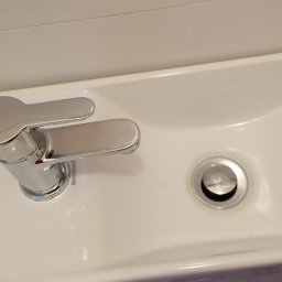 Magda Muszyńska - Biała, ceramiczna umywalka z chromowaną baterią Armatura i odpływem, zamontowana w łazience z białymi płytkami.