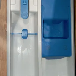 Szczegółowy widok szuflady dozownika pralki z resztkami detergentu i płynu do płukania, widoczne osady i zacieki na białym plastiku, niebieskie elementy dozownika.