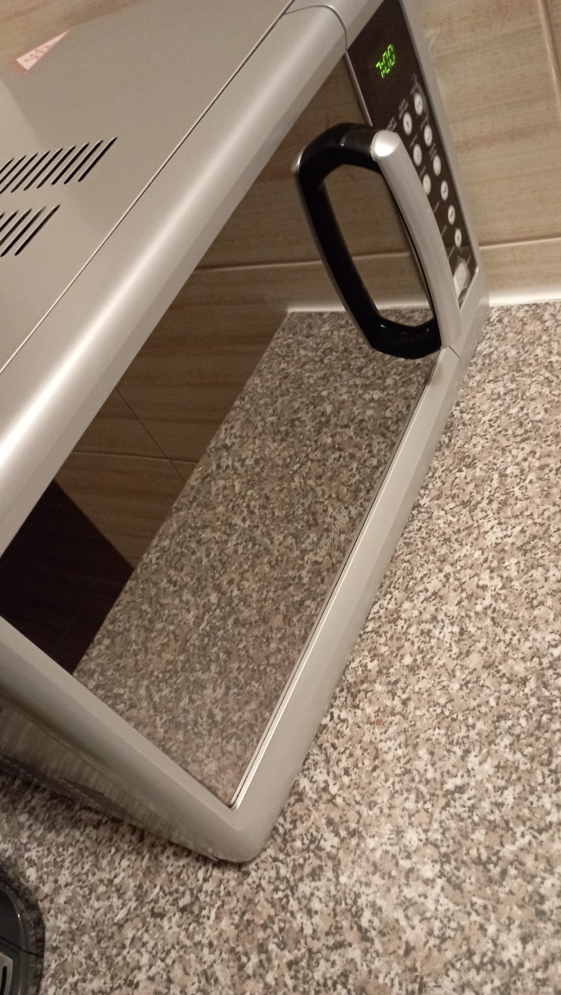 Błyszcząca, srebrna mikrofalówka na granitowym blacie, z odbiciem wnętrza i wyświetlaczem wskazującym godzinę 7:00.