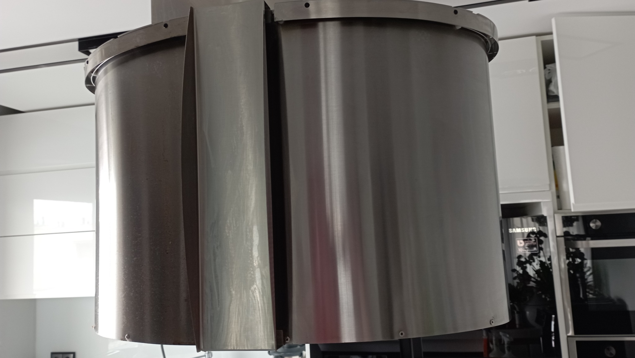 Cylindryczny okap kuchenny ze stali nierdzewnej, widoczne białe szafki kuchenne w tle, delikatne odbicie światła na powierzchni metalu.