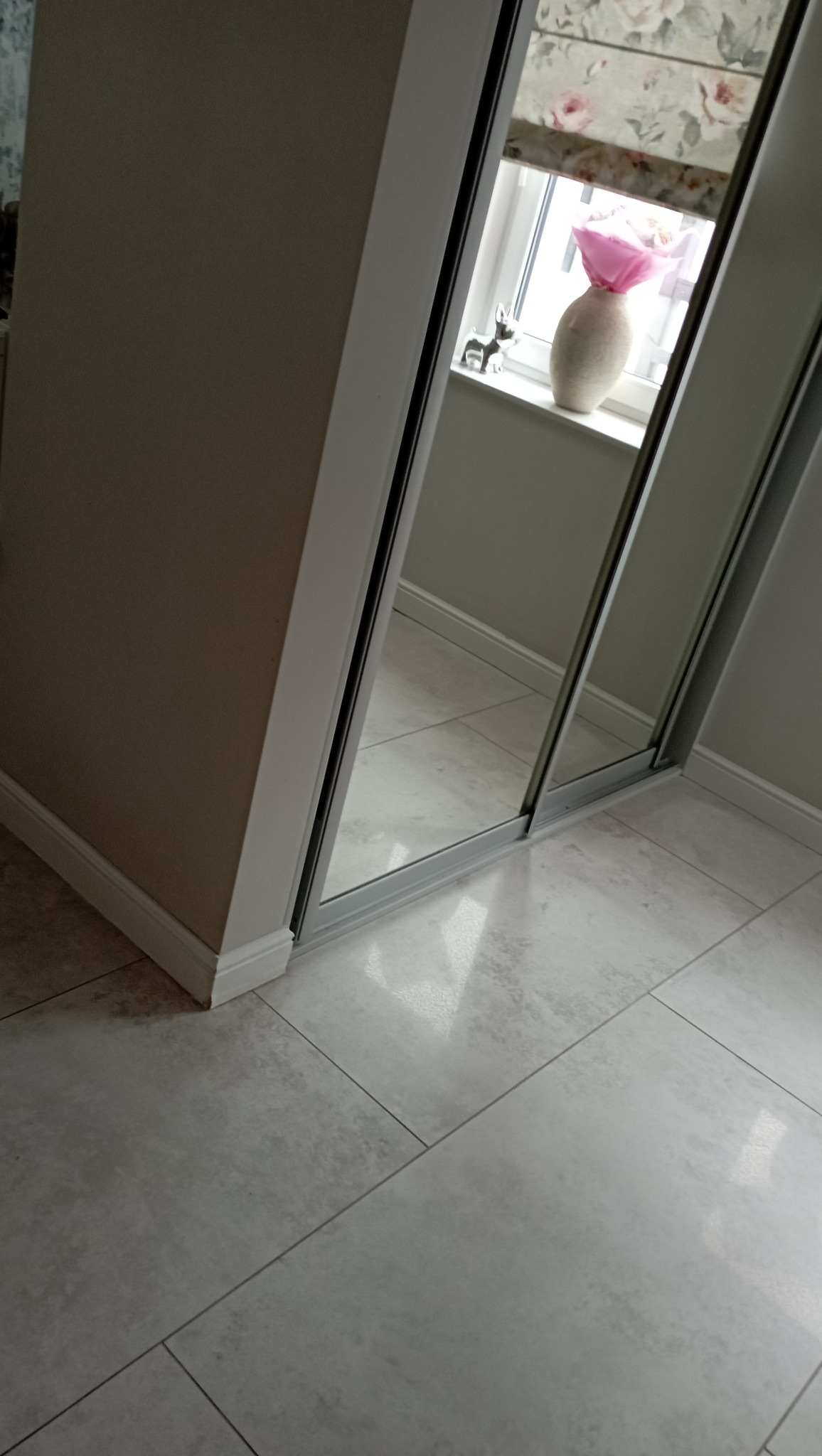Fragment szafy przesuwnej z lustrami, odbijającej okno z kwiatami w wazonie i figurką psa, na tle jasnej podłogi z płytek.
