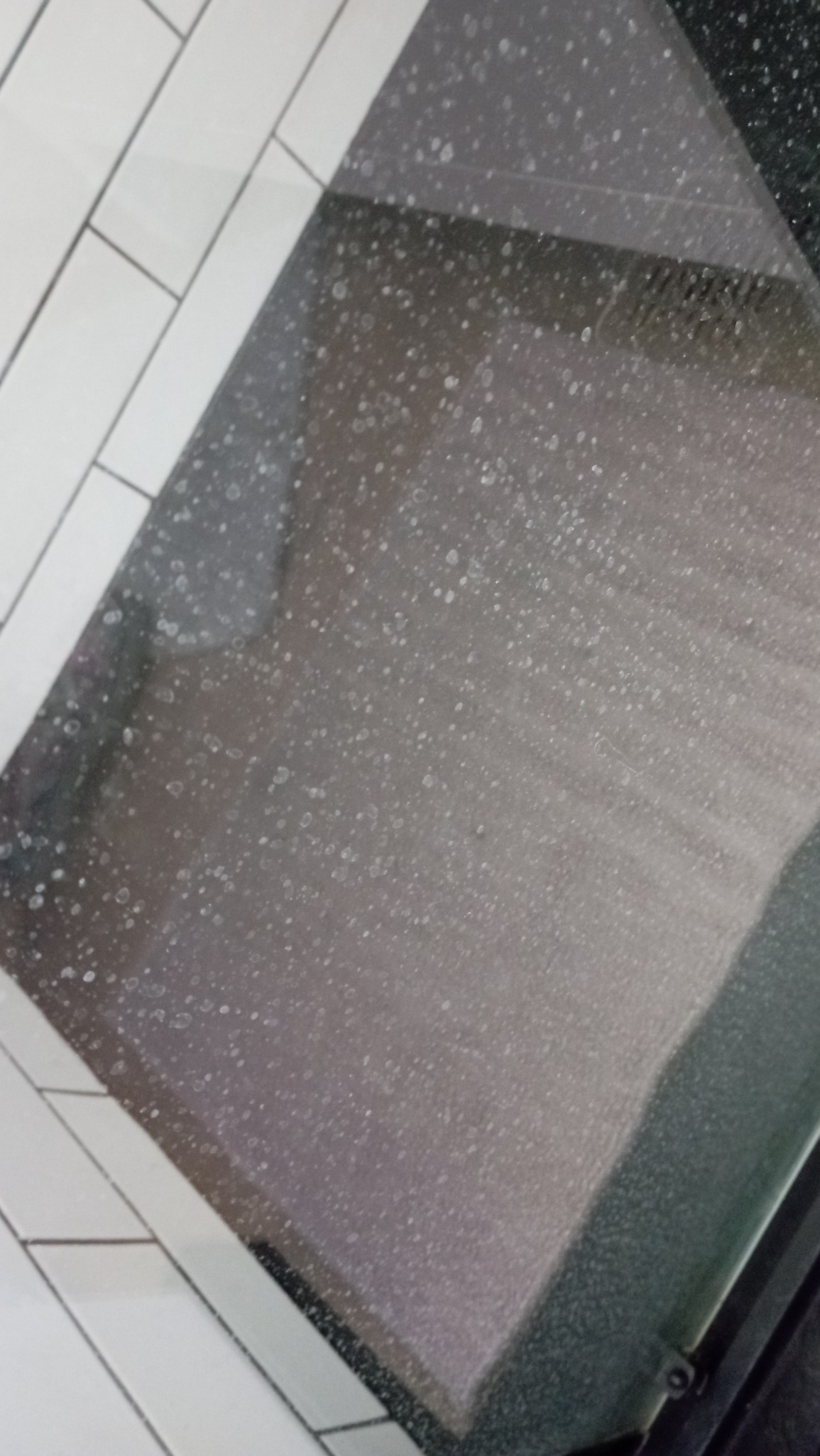 Brudna szyba piekarnika pokryta tłustymi plamami i osadem, widoczne białe płytki na ścianie w tle.