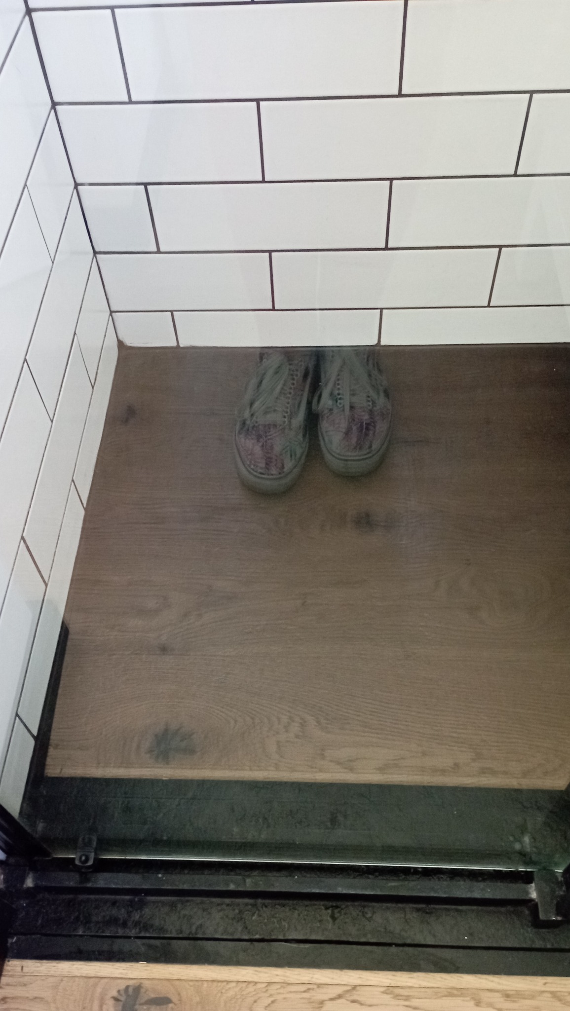 Para butów na drewnianej podłodze przy białych, kafelkowych ścianach z ciemnymi fugami, widoczne ślady użytkowania na podłodze.