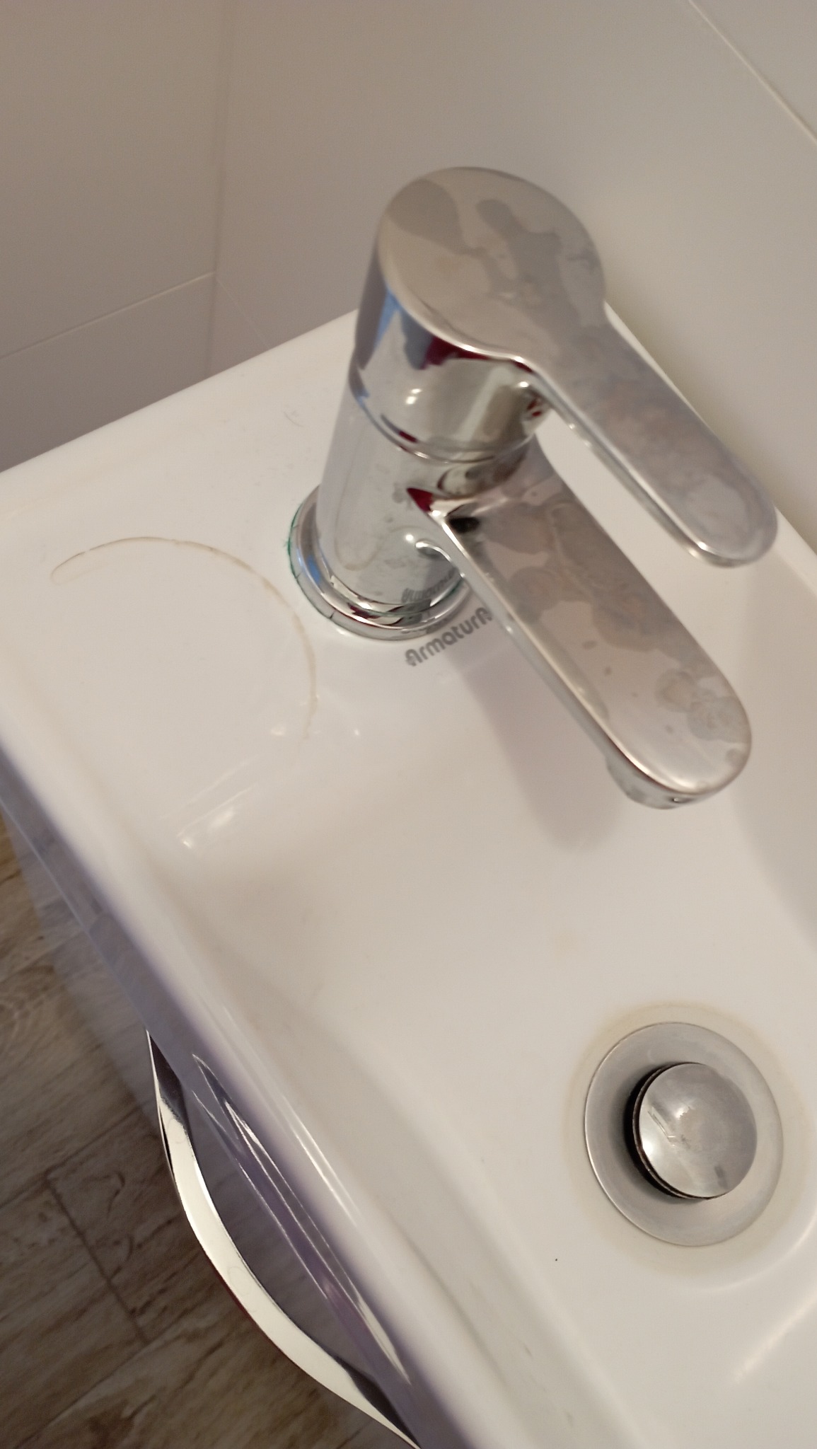 Umywalka z chromowaną baterią Armatura z widocznymi śladami po wodzie i zaciekami, odpływ umywalki widoczny w prawym dolnym rogu, ujęcie z góry.