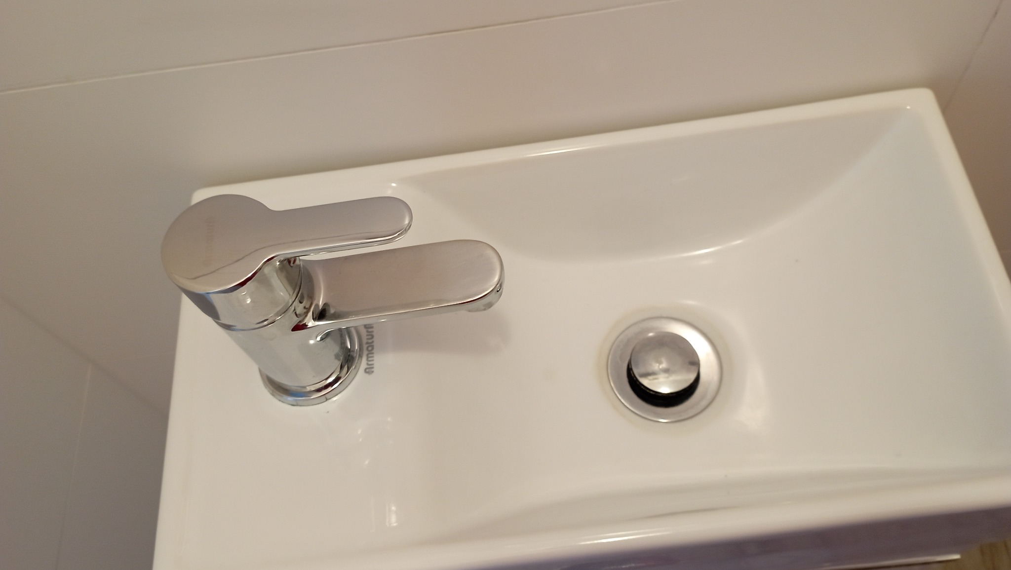 Biała, ceramiczna umywalka z chromowaną baterią Armatura i odpływem, zamontowana w łazience z białymi płytkami.