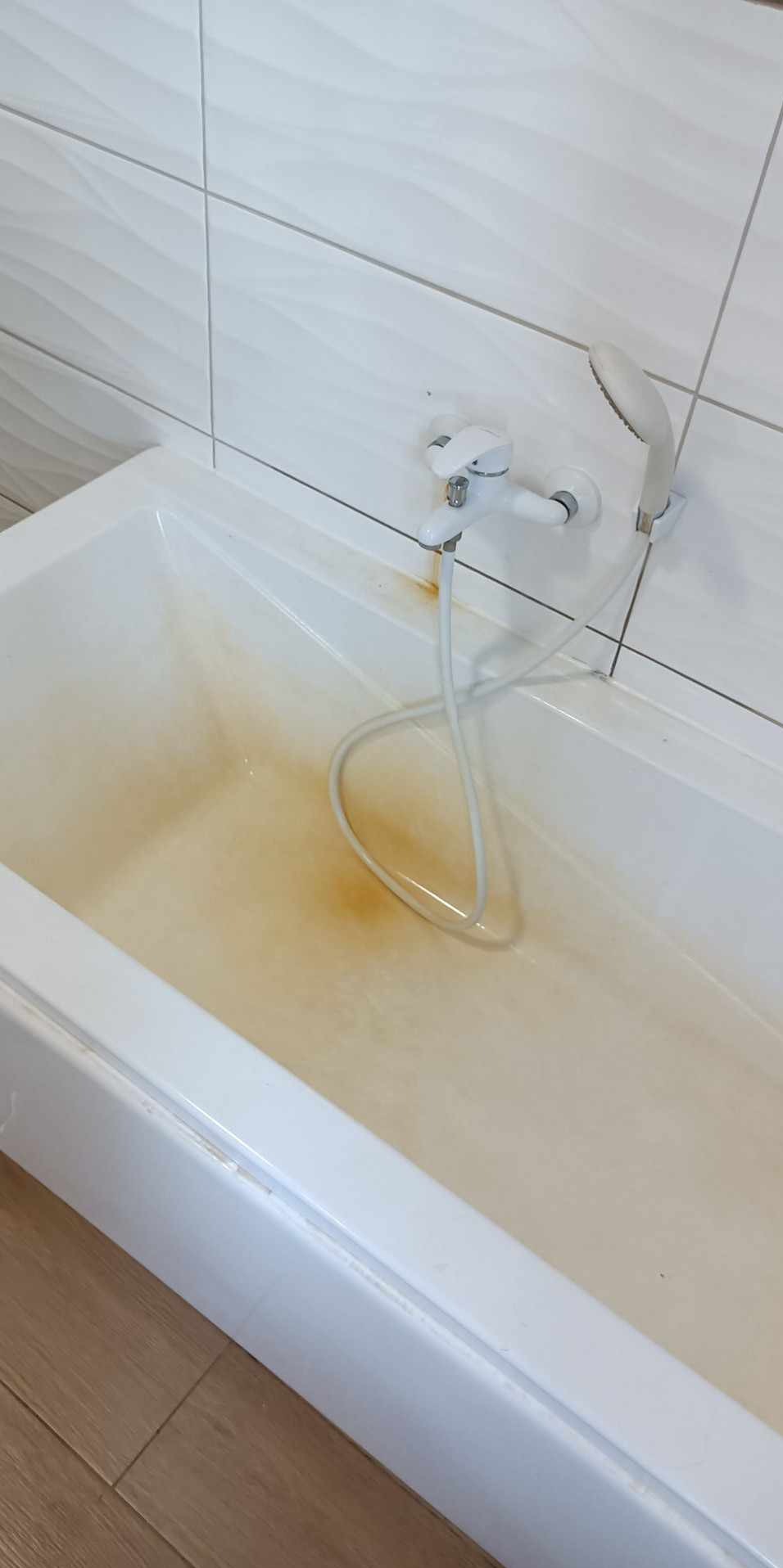 Biała wanna wypełniona brudną, rdzawą wodą. Widoczna biała armatura i fragment białych płytek na ścianie. Na dnie wanny widoczne osady i zacieki.