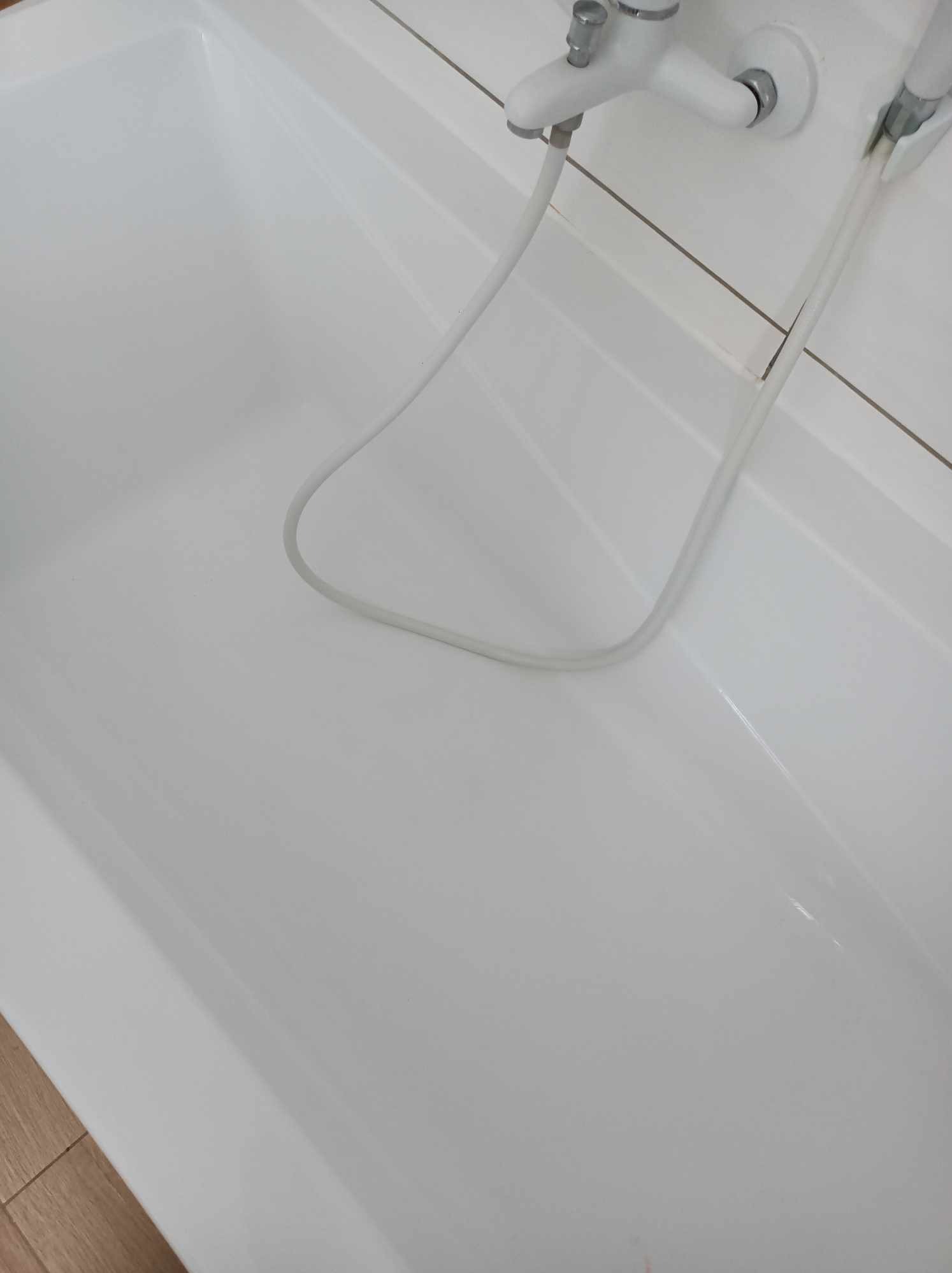 Biała, pusta wanna z baterią i słuchawką prysznicową. Widoczne białe płytki na ścianie i fragment drewnianej podłogi.