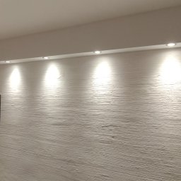 Krzysztof Zdun - Ściana z fakturą imitującą drewno pomalowana na biało, oświetlona przez sześć punktowych lamp sufitowych; na pierwszym planie fragment ciemnego mebla i papierowa torba.