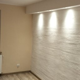 Krzysztof Zdun - Pusty pokój po remoncie z panelami podłogowymi w odcieniach brązu i beżu, pomalowaną na beżowo ścianą oraz ścianą z dekoracyjną strukturą podświetloną punktowymi lampami sufitowymi.