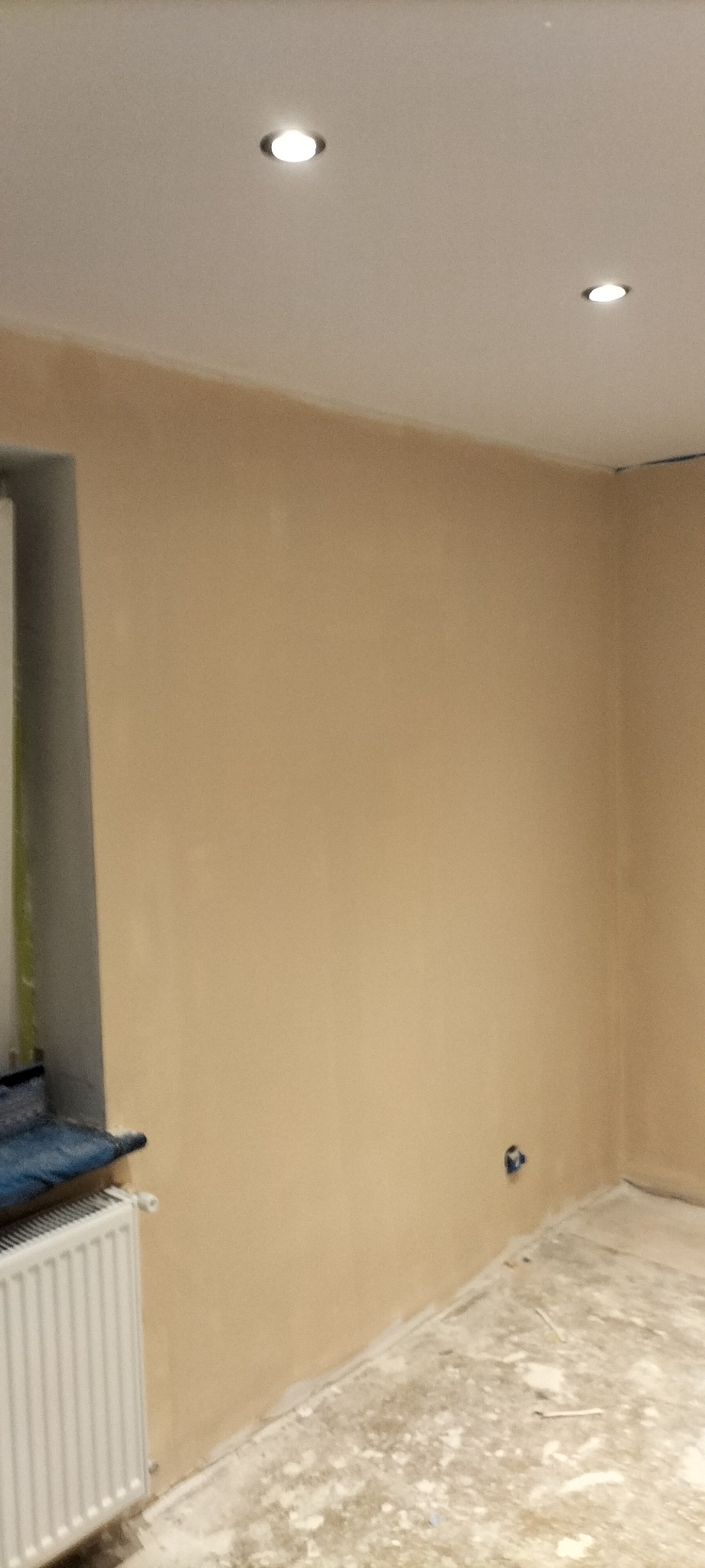 Ściana po szpachlowaniu, widoczne nierówności i przygotowanie do dalszych prac wykończeniowych, dwa halogeny w suficie, grzejnik pod oknem przykryty folią.