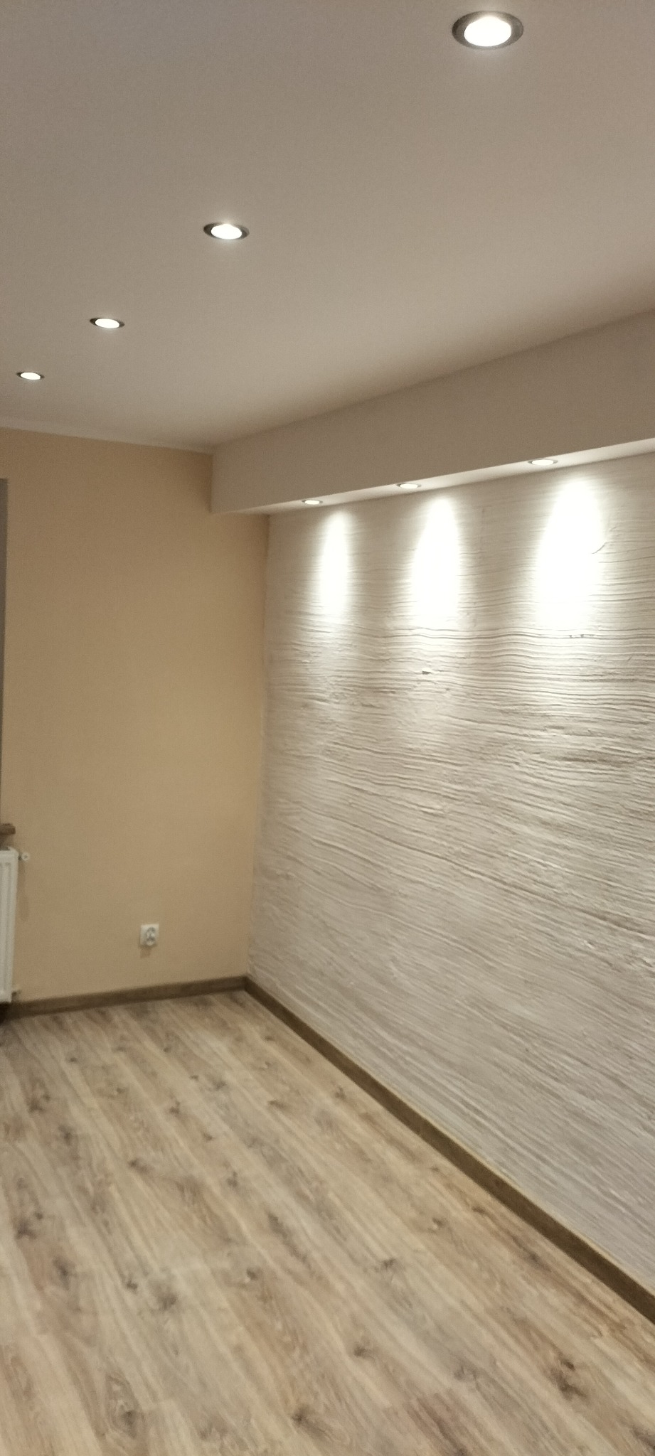 Pusty pokój po remoncie z panelami podłogowymi w odcieniach brązu i beżu, pomalowaną na beżowo ścianą oraz ścianą z dekoracyjną strukturą podświetloną punktowymi lampami sufitowymi.