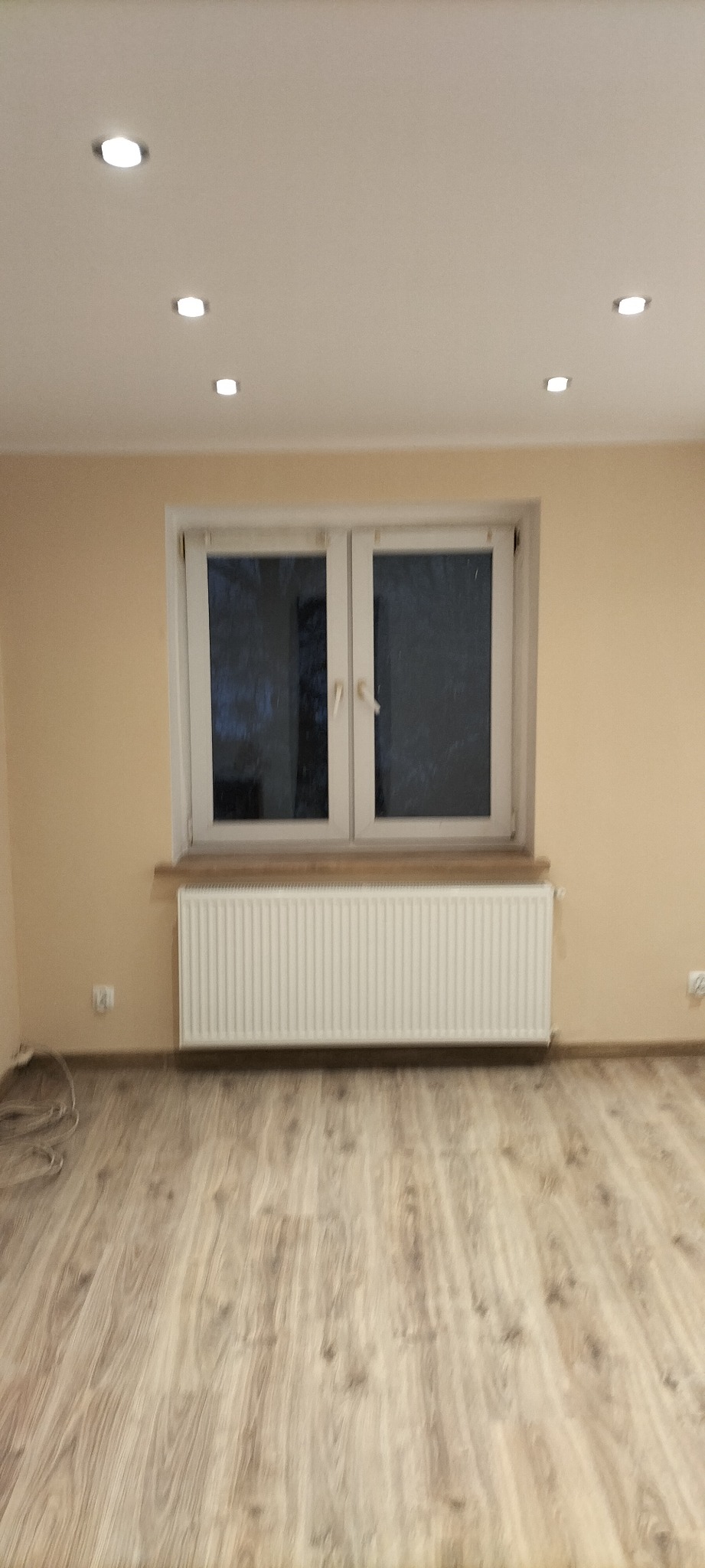 Jasny pokój po remoncie z panelami podłogowymi, białym grzejnikiem pod oknem i beżowymi ścianami z zamontowanym oświetleniem punktowym w suficie.