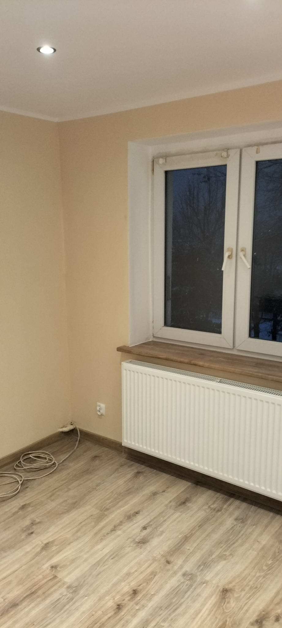 Pomalowane na beżowo wnętrze pokoju po remoncie, z widocznym grzejnikiem pod drewnianym parapetem, świeżo ułożonymi panelami podłogowymi i gniazdkiem elektrycznym przy podłodze.