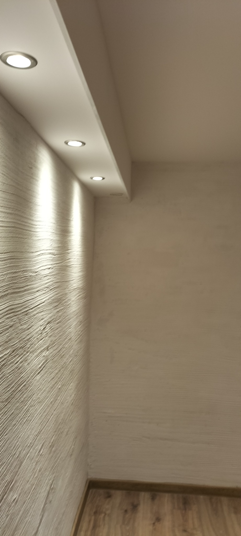 Wnętrze pokoju z oświetleniem punktowym wpuszczonym w podwieszany sufit, ściana z wyraźną fakturą i panele podłogowe w odcieniach brązu.