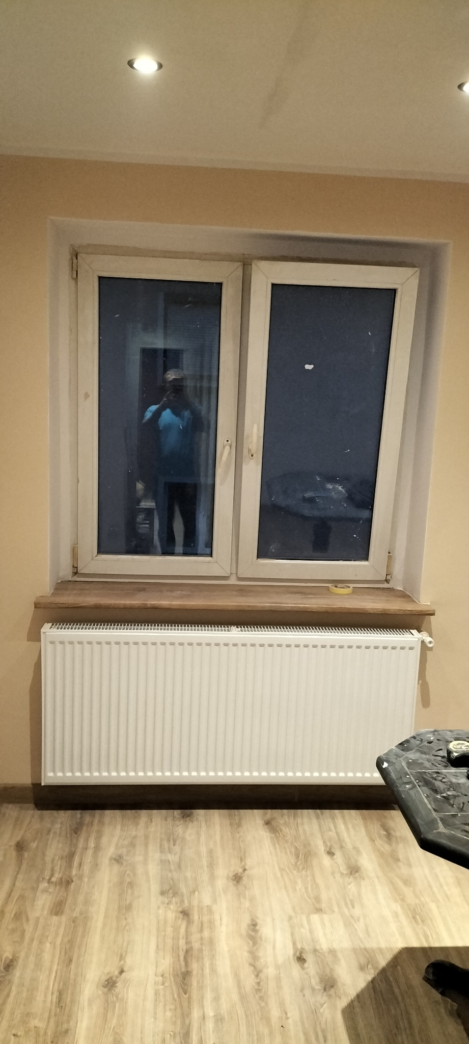 Biały grzejnik pod oknem z drewnianym parapetem, widoczna podłoga z paneli i odbicie osoby robiącej zdjęcie w szybie okiennej.