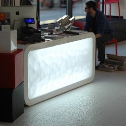 Łukasz Cendrowski - Nowoczesna lada recepcyjna z podświetlanym frontem o nieregularnej fakturze, z komputerem iMac, drukarką fiskalną i dekoracyjnym układem książek na blacie, w tle siedzący mężczyzna na czerwonym...