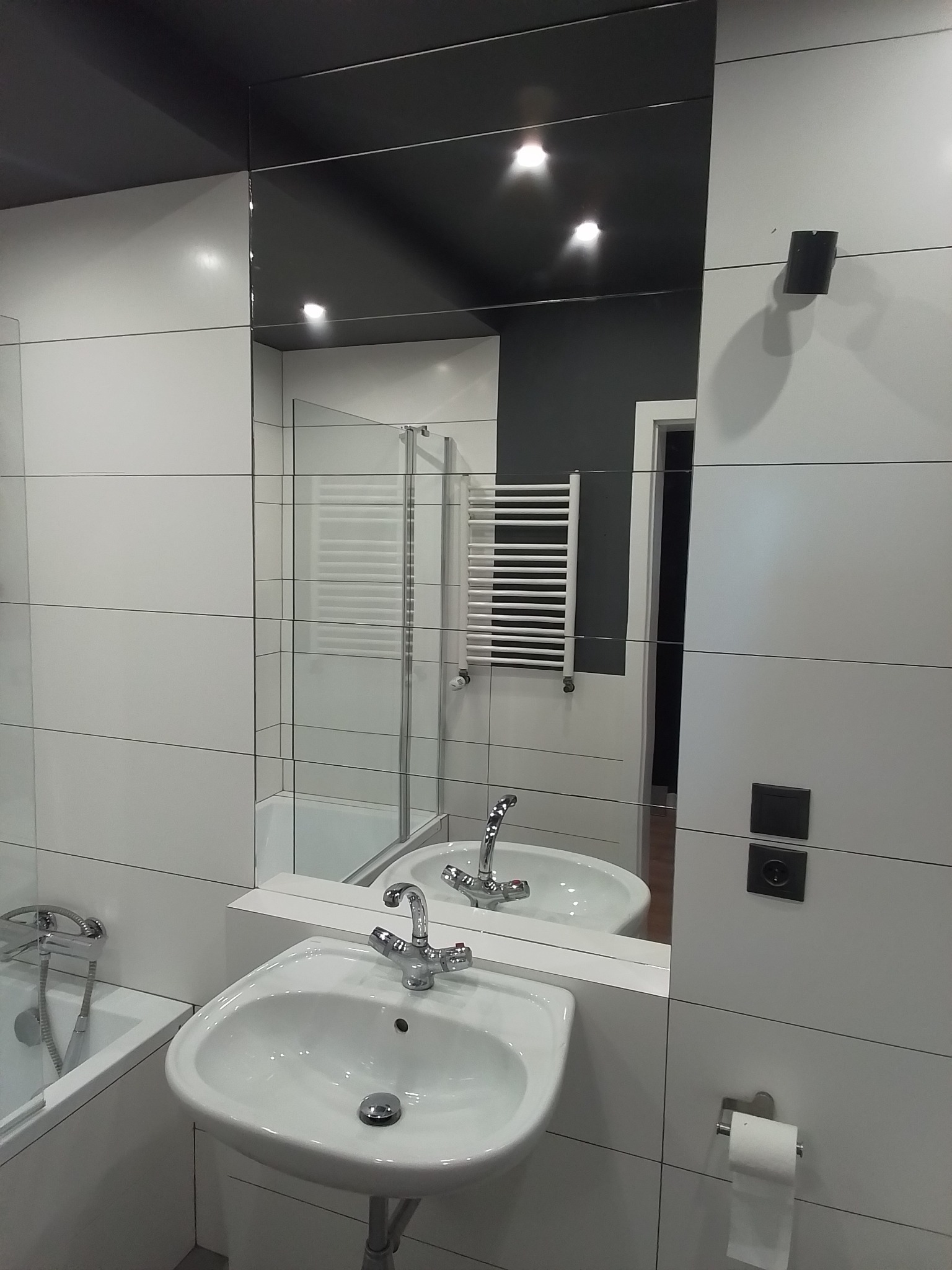 Nowoczesna łazienka z białymi, prostokątnymi płytkami, czarnym sufitem i dużym lustrem, w którym odbija się kabina prysznicowa i grzejnik drabinkowy.